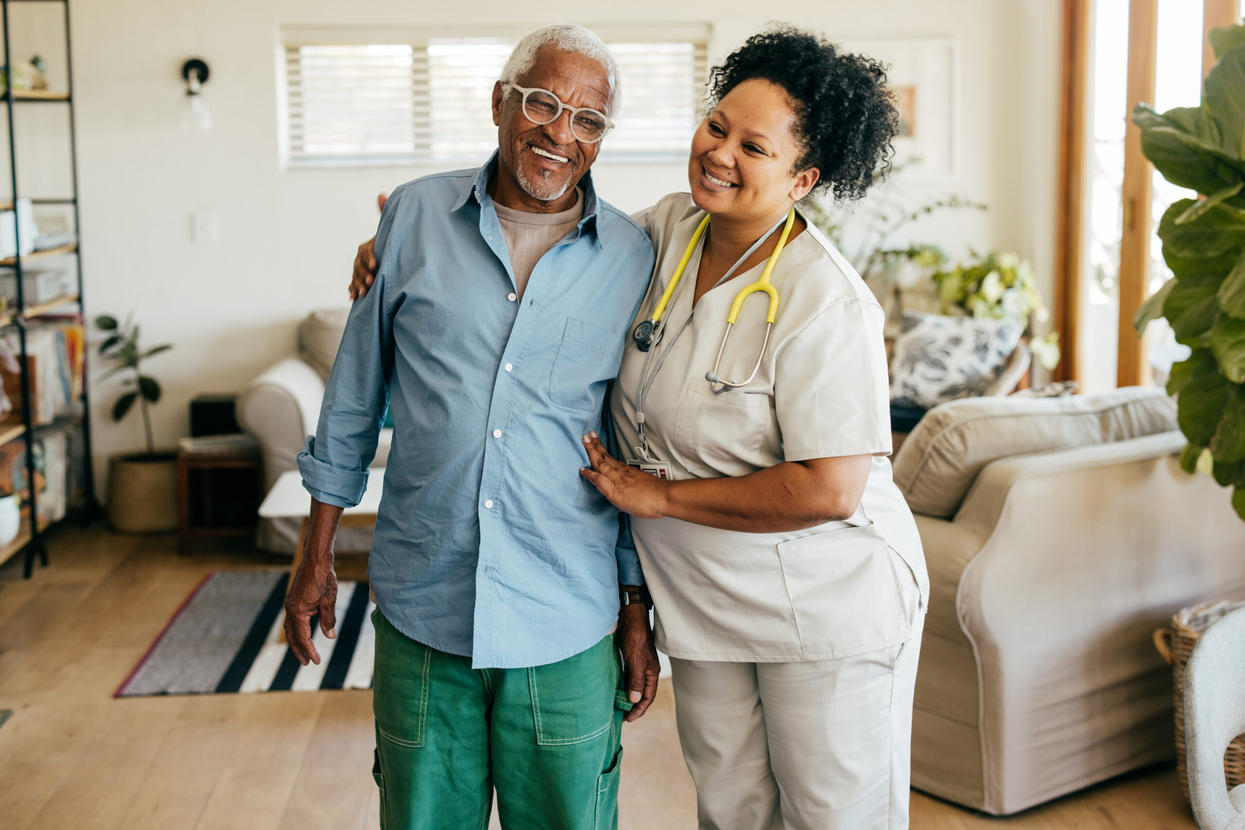 A nurse in scrubs holding up an old man and smiling at the camera.